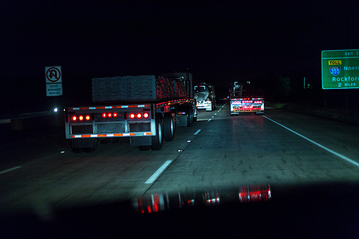 Car driver's point of view following three large trailer trucks speeding along a multiple lane expressway highway at night somewhere in the state of Illinois, USA.