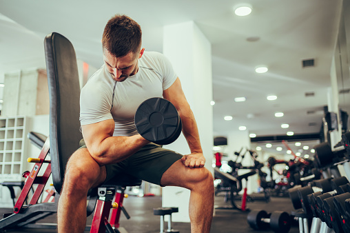 A strong muscular sportsman is sitting in a gym during his strength training and pumping muscles while looking at it. A dedicated sportsman is sitting in a gym and doing exercises for the biceps.