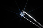 Diamond selective focus held in metal jeweller tweezers, brilliant stone cut with star shape light ray burst, starburst or sunbeam. Inspection of polish quality and contamination. Carbon material.