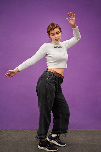 Stylish young woman dancing with her hands in the air in front of a purple wall on a city sidewalk