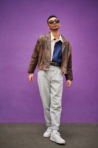 Portrait of a young man wearing stylish sunglasses posing in front of a purple wall on a city sidewalk