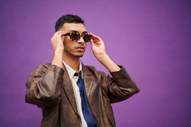 Young man putting on stylish sunglasses against a purple background