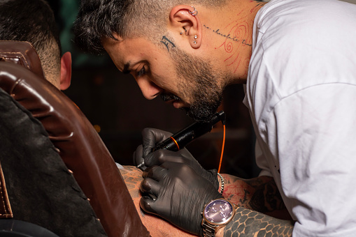 Tattooer master's hand in black glove making tattoo art on male hand with machine for tattoo over dark background.