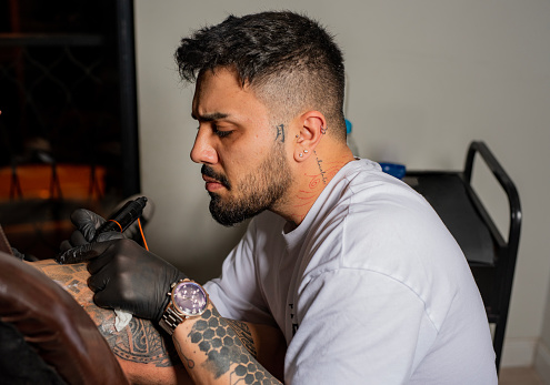 Tattooer master's hand in black glove making tattoo art on male hand with machine for tattoo over dark background.