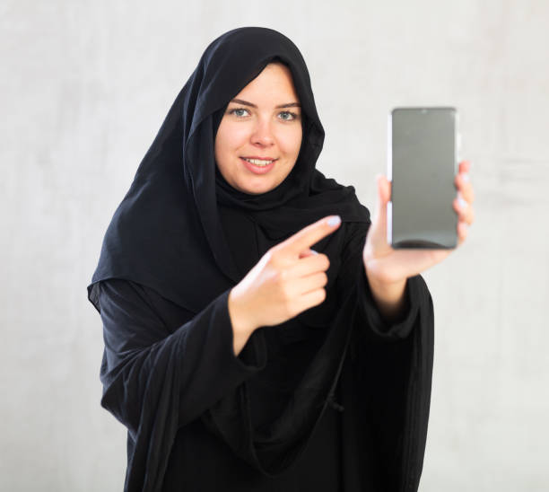 smiling Muslim woman in traditional black hijab shows blank smartphone screen close-up young woman in hijab shows dark empty smartphone screen. Close-up, gray background azerbaijani culture stock pictures, royalty-free photos & images