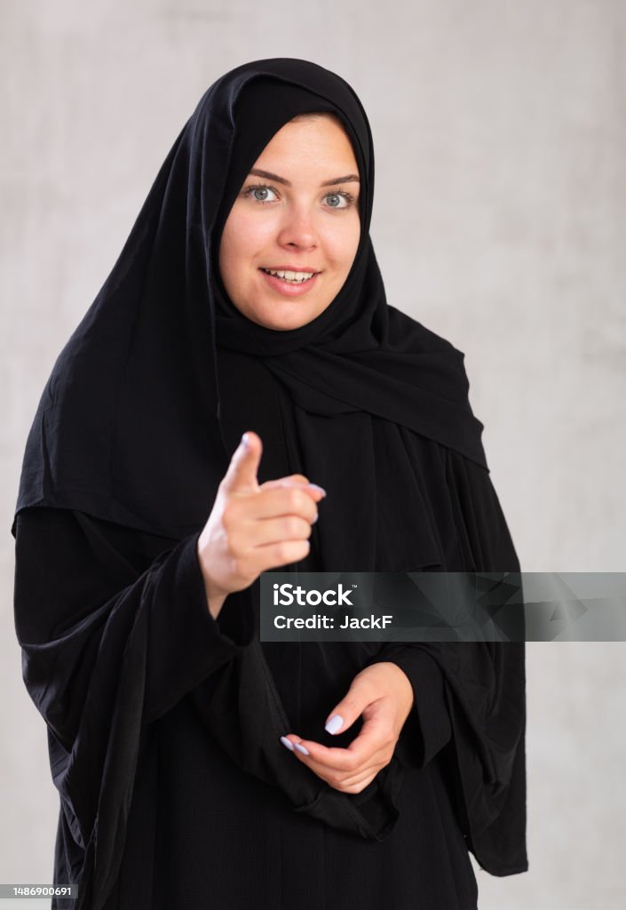 Young female in black hijab pointing at you - Portrait of young woman pointing her finger woman in black hijab points her hand to side.positive Muslim woman raised her index finger and urges to be attentive. Close-up, gray background 20-24 Years Stock Photo