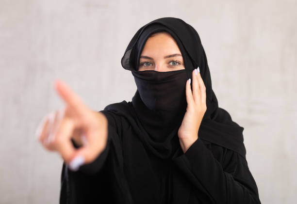 woman with her face covered by burka points her hand to side woman with her face covered by burka points her hand to side. Positive Muslim woman raised her index finger and urges to be attentive. Close-up, gray background azerbaijani culture stock pictures, royalty-free photos & images