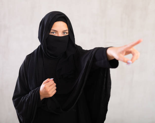 woman with her face covered by burka points her hand to side woman with her face covered by burka points her hand to side. Positive Muslim woman raised her index finger and urges to be attentive. Close-up, gray background azerbaijani culture stock pictures, royalty-free photos & images