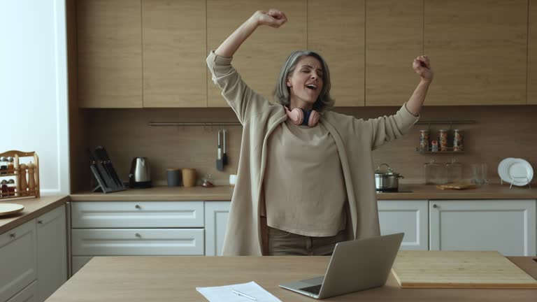 Mature woman distracted from working on laptop, dancing in kitchen