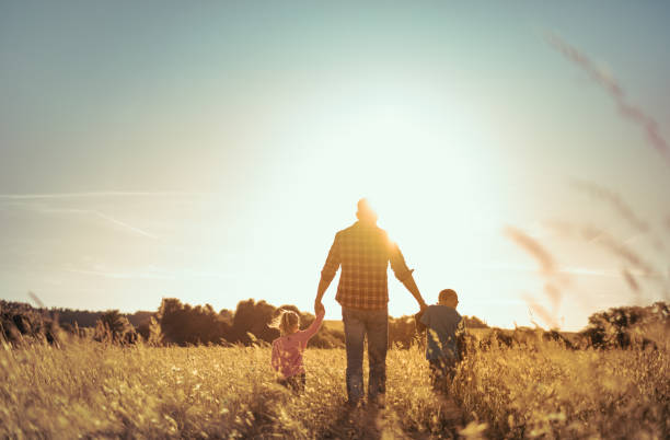 Vater und Kinder, die bei Sonnenuntergang Händchen halten, gehen auf einer Wiese spazieren – Foto