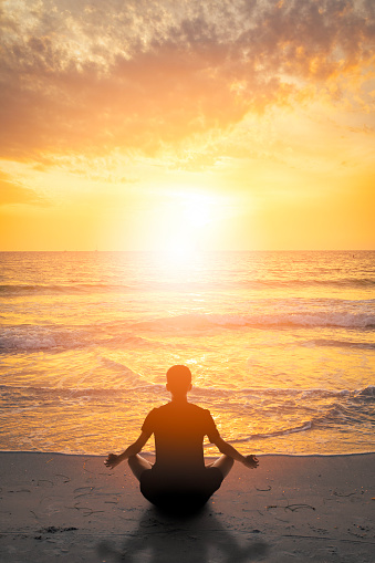 A young man sitting in a meditative position on the beach during sunset.
