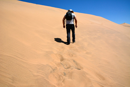 A tired man with a backpack on his back and wearing a cap climbs a desert sand hill. Rear view. Unknown person