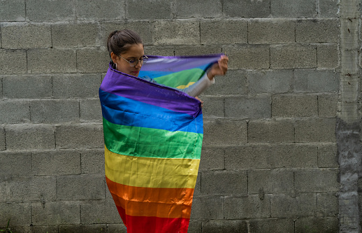 outdoor woman draped with gay pride flag waving it on gray background, portrait of young woman on gray brick background with gay pride flag