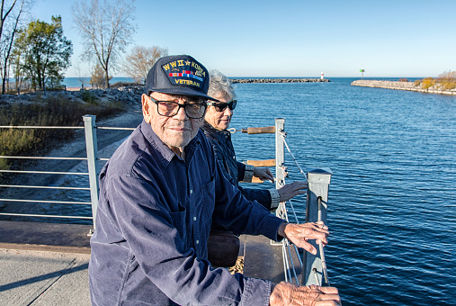 An aging but still active 93 year old World War II and Korean Conflict USA military war veteran and his senior adult woman daughter are sightseeing - standing together side by side at Lake Ontario near Rochester, New York State, USA. He is looking at the camera curiously.