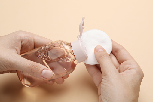 Woman pouring micellar water from bottle onto cotton pad against beige background, closeup