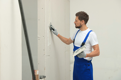Professional worker in uniform plastering wall with putty knife indoors