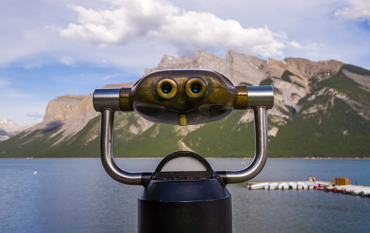 Coin operated binoculars on Lake Minnewanka. Rocky mountains, clear blue water and green forest. Summer vacation in the mountains - a trip to Banff, Alberta, Canada