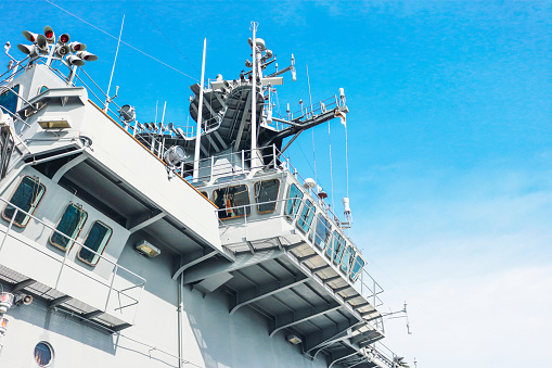 One crew member is raising two maritime flags, while the other watches or assists, on the bridge of USS Canberra (LCS 30), an Independence Class combat ship of the US Navy which is docked at Garden Island in Sydney Harbour.  She was in port for her commissioning ceremony on 22 July 2023.  As part of the ceremony, her captain was presented with a kangaroo insignia attached to all ships of the Royal Australian Navy, but decorated in the colours and design of the Stars and Stripes.  That insignia has been attached to the ship and visible on the right.  The name of the ship and serial number is visible on the orange life ring.  A large, padded chair is visible on the bridge.  This image was taken on a cloudy afternoon on 29 July 2023.