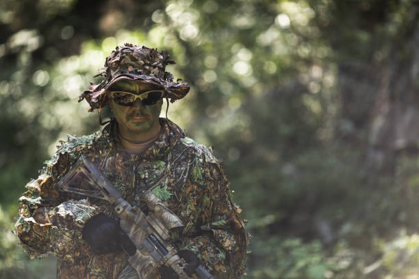 A man in a 3D leaf style light weight Ghillie suit and face paint camouflage with an AR-15 rifle with ACOG scope in the woods.
