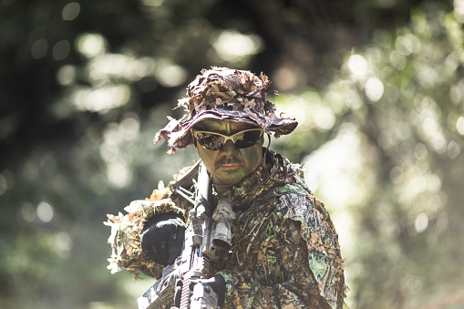 A man in a 3D leaf style light weight Ghillie suit and face paint camouflage with an AR-15 rifle with ACOG scope in the woods.