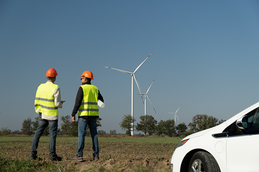 Windmills generate energy for electric cars in countryside. Wind turbine technicians in orange helmets stand against powerful windmills