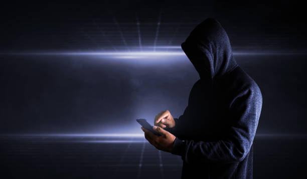 hacker usando smartphone. hombres vestidos de negro con la cara oculta miran el teléfono inteligente - business stealing top secret confidential fotografías e imágenes de stock