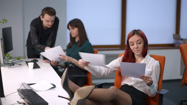Careless young woman with feet on table throwing around paperwork with busy blurred colleagues talking at background. Portrait of carefree Caucasian female employee in office with coworkers.