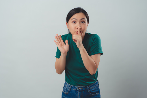 Asian woman happy smiling putting index finger on lips meaning keeping secret, standing isolated on background.