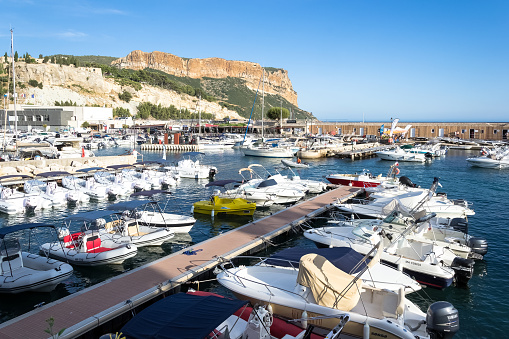 Cassis, France - August 2019 - View of the harbor of Cassis, a charming commune located east of Marseille in the southern region of France. \nSituated on the French Riviera, Cassis is a popular tourist destination renowned for its picturesque coastline, stunning beaches, and charming village atmosphere. The harbor is a hub of activity, offering visitors the chance to admire colorful fishing boats, dine at seafood restaurants, and stroll along the promenade.