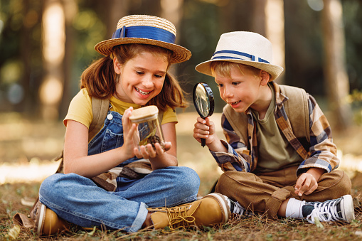 Two little children boy and girl with backpacks looking examining plants and insects in a jar through magnifying glass while exploring forest nature and environment on sunny day during outdoor ecology school lesson