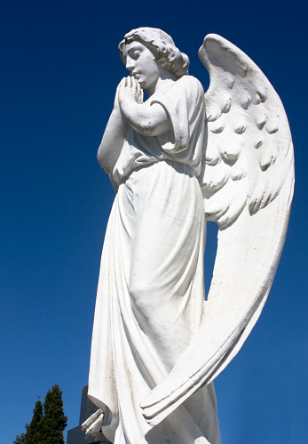 Ancient white marble angel sculpture,  seen on tombstone in a graveyard, low angel view, Lugo city, Galicia, Spain. Clear blue sky background.
