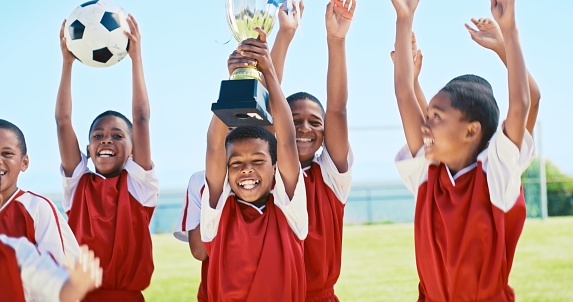 Trophy, football and winning children celebrate on field for achievement, teamwork or victory. Soccer, celebration award or happiness of kids with sports success, team reward or competition champions