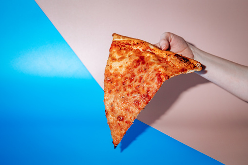 Hand holding slice of pizza in front of blue and pink background