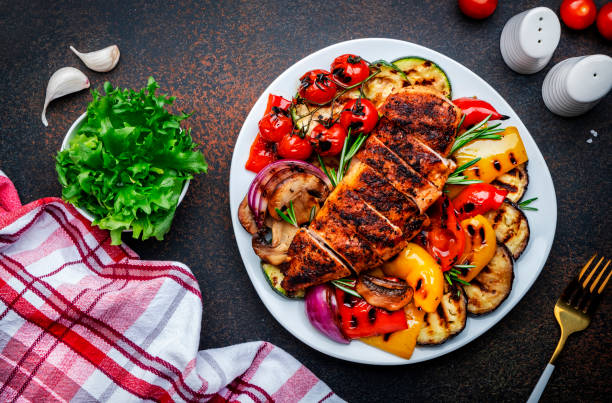 Grilled vegetables and chicken fillet salad. Paprika, zucchini, eggplant, mushrooms, tomatoes, onion and rosemary, served on plate, brown table background, top view stock photo