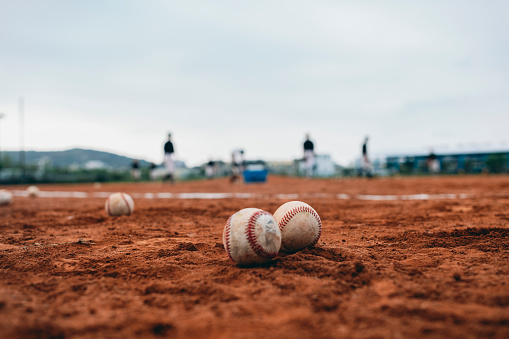 Baseballs scattered on the baseball field, with a blurry background of a baseball team practicing in the distance.
