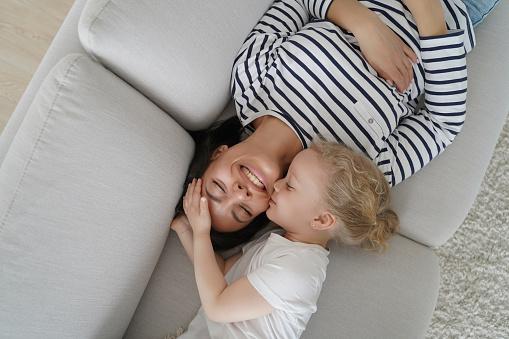 Happy mom having rest with her little daughter on couch. Family playing and relaxing together. Woman and kid are smiling and falling asleep together. Caucasian mother and child having daytime sleep.