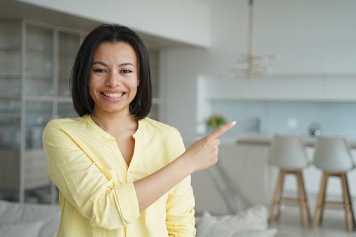 Smiling female real estate agent homeowner or tenant pointing with finger at copy space aside standing in new home. Happy positive woman recommend shopping offer, advertise service, looking at camera.