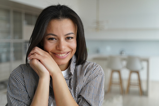 Smiling young female homeowner or apartment renter pose looking at camera at home. Happy shy brunette woman feels tender emotion, folded hands near cheek in new house. Mortgage, real estate sale.