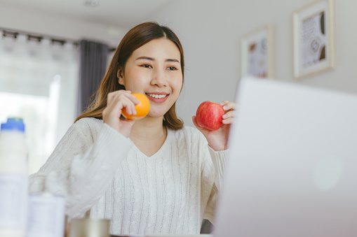 Young woman holding apple and orange and using a laptop.