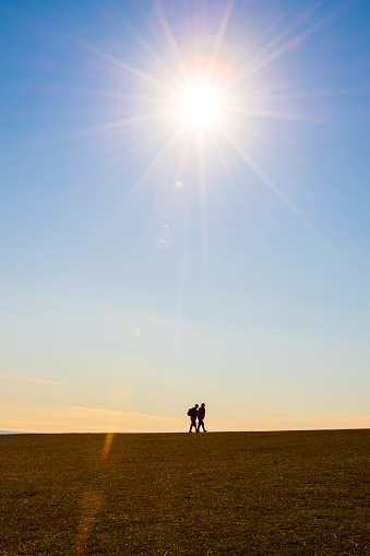 Two men in silhouette walk under the sun shining above a field.