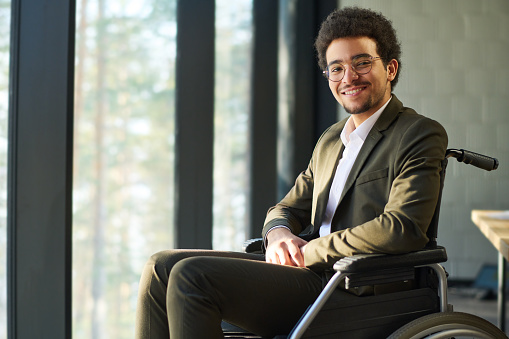 Happy young multi-ethnic businessman with disability sitting in wheelchair by large office window and looking at camera with smile