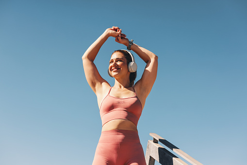 Woman in sportswear stretching her arms outdoors. Happy sportswoman doing a warm up exercise with headphones on. Female athlete working out against the sky.
