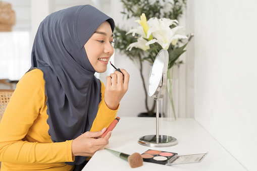 Asian young Muslim woman applying makeup lipstick on lips while looking in mirror