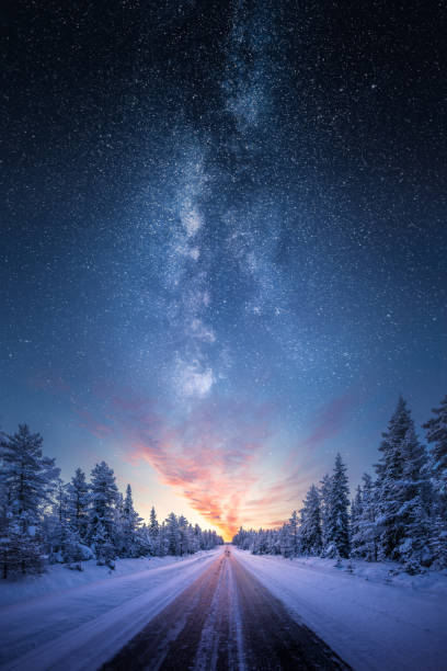 Road leading towards colorful sunrise between snow covered trees with epic milky way on the sky stock photo