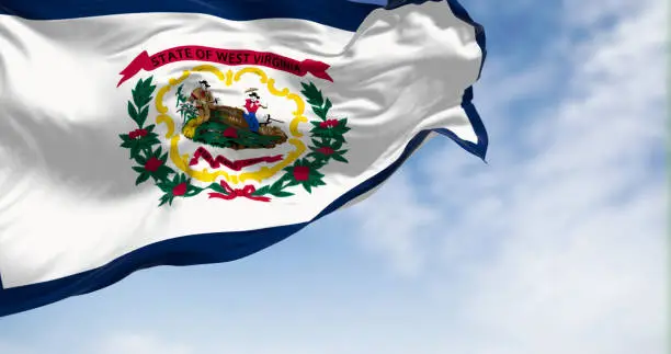 West Virginia state flag waving on the wind on a clear day. White field, blue stripe, state coat of arms, Rhododendron maximum, and red ribbon. 3d illustration render. Fluttering fabric.