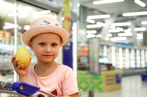 Portrait of funny cute little girl with lemon at food showcase store background, looking at camera. Pretty kid in pink t-shirt buying in big superstore. Shopping and purchase concept. Copy text space