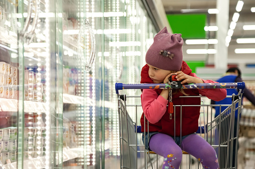 Pensive little girl sitting in shopping cart near showcase window in food store, looking away. Bored kid in red casual clothe buy retail in supermarket. Shopping and buying concept. Copy ad text space