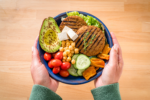 Overhead view of woman's hands holding a plate filled with healthy plant-based food. The composition include, tofu, cherry tomatoes, chickpeas, cucumber, lettuce, dried fruits, avocado and veggy patties. High resolution 42Mp studio digital capture taken with SONY A7rII and Zeiss Batis 40mm F2.0 CF lens