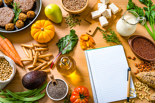 Overhead view of a wooden table filled with a large number of healthy plant-based food. A blank note pad with copy space is at the bottom left. The composition includes soy beans, soy milk, tofu, legumes, leaf vegetables, root vegetables, wholegrain pasta, avocado, chia seeds and veggy patties and meatballs in a cooking pan. High resolution 42Mp studio digital capture taken with SONY A7rII and Zeiss Batis 40mm F2.0 CF lens