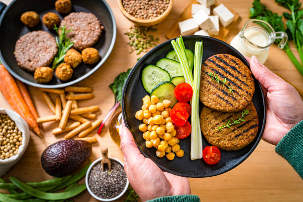 Overhead view of woman's hands holding a plate filled with healthy plant-based food. The composition includes soy beans, soy milk, tofu, legumes, leaf vegetables, root vegetables, wholegrain pasta, avocado, chia seeds and veggy patties and meatballs in a cooking pan. High resolution 42Mp studio digital capture taken with SONY A7rII and Zeiss Batis 40mm F2.0 CF lens
